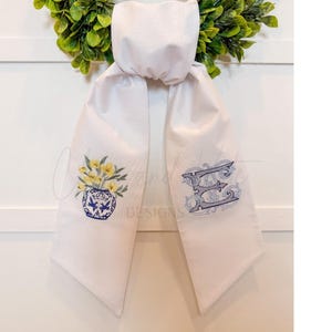 May include: White scarf with embroidered details. One end features a blue and white vase with yellow flowers. The other end displays an ornate blue letter "E". The scarf is hanging against a white background with a green wreath.