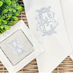 May include: White linen coaster and napkin set with light blue embroidered monogram. The coaster has a black stitched border. The napkin and coaster feature the same elegant monogram design. The set is displayed on a woven surface with greenery.