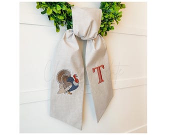 May include: A gray fabric bow with a turkey and the letter "T" embroidered on it. The bow is tied around a green wreath.