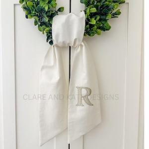 May include: A white satin door bow with the letter "R" embroidered in gold thread. The bow is tied around a green wreath.