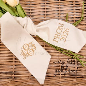May include: Ivory fabric sash with gold thread embroidery. Features a monogram and the text "PROM 2026". The sash is displayed on a woven wicker surface with a bouquet of yellow flowers.