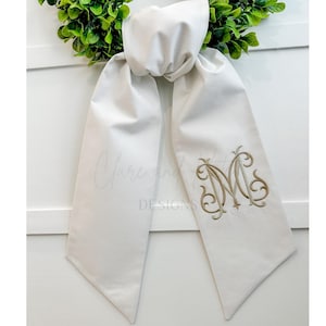 May include: Cream-colored scarf with an embroidered gold monogram. The scarf is knotted at the top and hangs down with pointed ends. The text "Clare and Designs" is visible on the scarf.