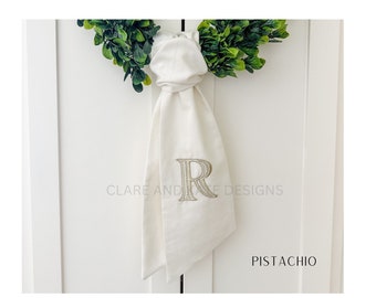 May include: A white fabric towel featuring an embroidered letter R in a light green thread color. The towel is knotted at the top and hangs below a green leafy wreath. The text CLARE AND KATE DESIGNS and PISTACHIO are visible on the image.