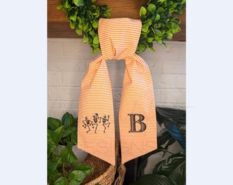 May include: A long, orange and white gingham fabric banner featuring black embroidery. The left side displays three dancing skeletons, while the right side features a large, stylized capital letter B in a bold, serif font.