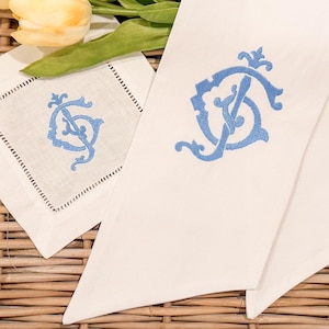 May include: White linen napkin and coaster set with light blue embroidered monogram. The napkin is folded, and the coaster is square with a decorative stitched border. Yellow tulips and greenery are in the background.