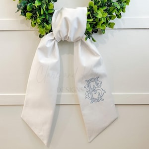 May include: A white fabric wreath sash with a light blue embroidered monogram. The sash is draped over a green wreath and white wall. The monogram features the letters "S" and "G" intertwined in an elegant script.