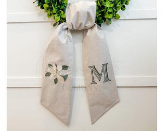 May include: A beige fabric wreath sash with a magnolia flower and the letter "M" embroidered in dark green. The sash is hanging on a white background with a green wreath.