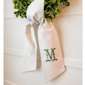 May include: A white fabric wreath bow with a green embroidered monogram "M" on the front.