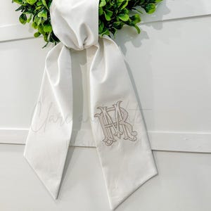 May include: White fabric sash with a monogram of the letters "HR" embroidered in a gray color.