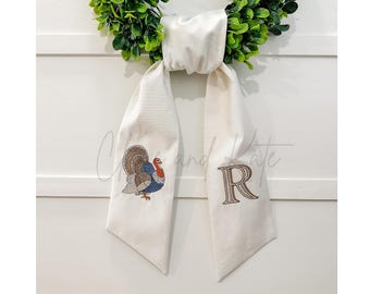 May include: Cream-colored Thanksgiving scarf with a detailed embroidered turkey and the letter "R". The scarf is draped over a green wreath, creating a festive and decorative display.