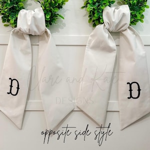 May include: Two white fabric door swags with a black embroidered letter "D" and green wreath accents. The words "opposite side style" are written in black script below the swags.
