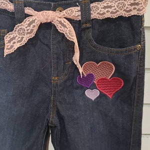 May include: Dark blue denim jeans with a pink lace belt and a patch of four embroidered hearts in shades of red, purple, and pink.