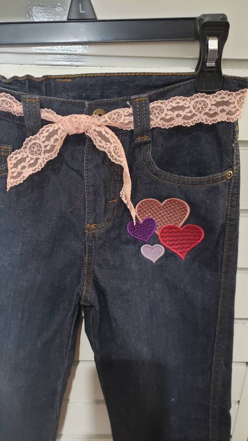 May include: Dark blue denim jeans with a pink lace belt and a patch of four embroidered hearts in shades of red, purple, and pink.