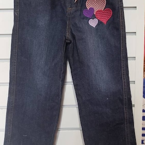 May include: Dark blue denim capri pants with a pink lace belt and a multi-coloured heart applique on the right leg.