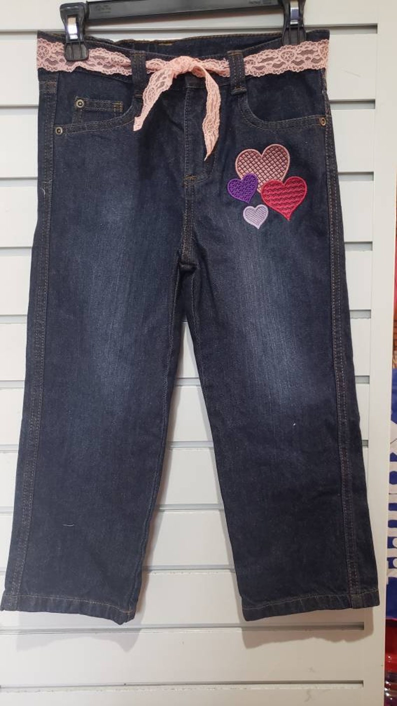 May include: Dark blue denim capri pants with a pink lace belt and a multi-coloured heart applique on the right leg.