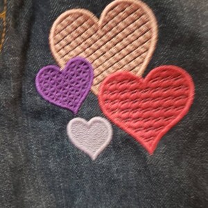 May include: Four embroidered hearts on a dark blue denim fabric. The largest heart is pink, the next largest is red, the next is purple, and the smallest is light purple.