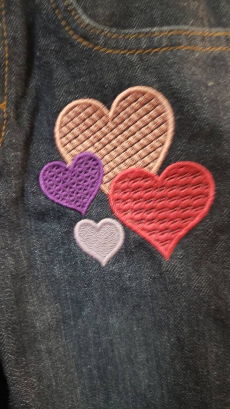 May include: Four embroidered hearts on a dark blue denim fabric. The largest heart is pink, the next largest is red, the next is purple, and the smallest is light purple.