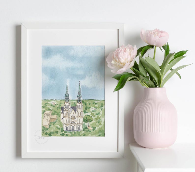 May include: A watercolor painting of a church with two tall spires, surrounded by green trees and a blue sky with white clouds. The painting is framed in a white frame and is displayed on a white wall.