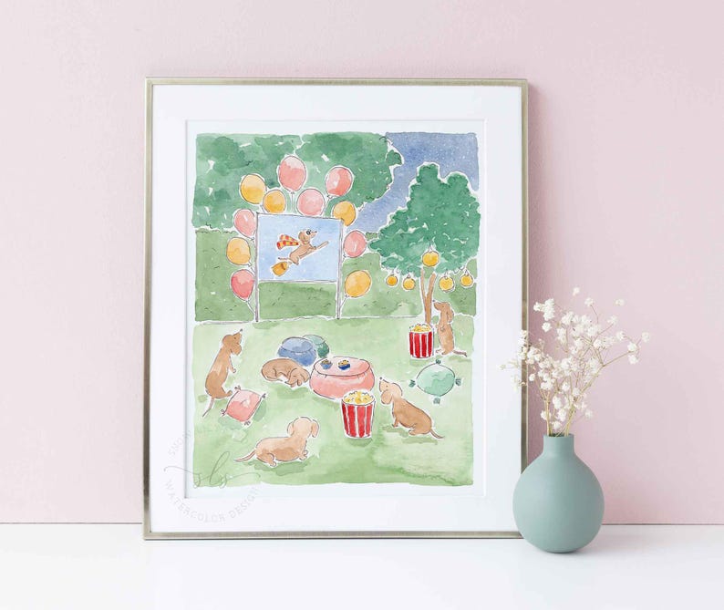 May include: Watercolour print depicting a dog party. The artwork shows dachshunds, balloons, popcorn, and a dog-themed movie screen. The scene is set in a park with a green lawn and trees. The print is framed in a silver frame.
