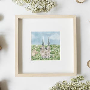May include: A framed watercolor painting of a church with two tall spires. The church is painted in shades of white, gray, and green, and is set against a blue sky with white clouds. The painting is surrounded by a white mat and a light wood frame.