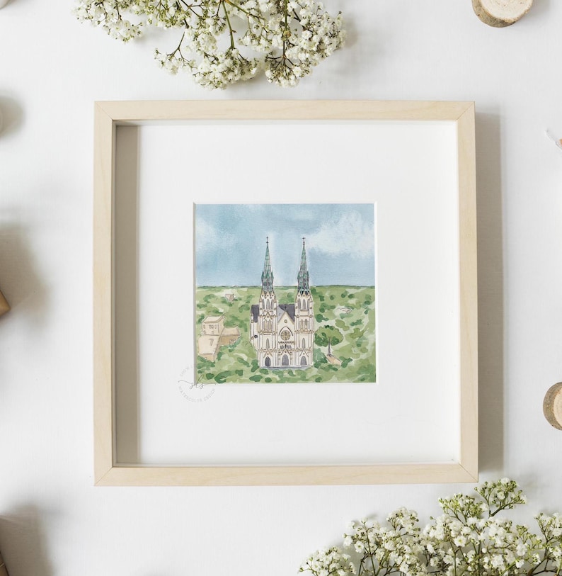 May include: A framed watercolor painting of a church with two tall spires. The church is painted in shades of white, gray, and green, and is set against a blue sky with white clouds. The painting is surrounded by a white mat and a light wood frame.