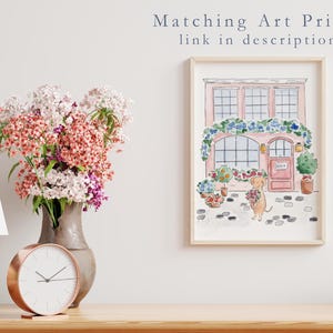 May include: A framed watercolour print of a pink building with floral decorations and a dog holding flowers. A vase of flowers, a lamp, and a clock are on a wooden surface. Text above the print reads "Matching Art Prints link in description."