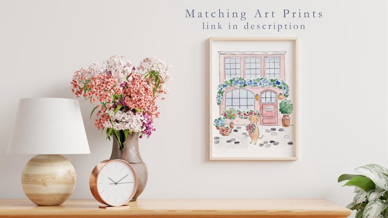 May include: A framed watercolour print of a pink building with floral decorations and a dog holding flowers. A vase of flowers, a lamp, and a clock are on a wooden surface. Text above the print reads "Matching Art Prints link in description."