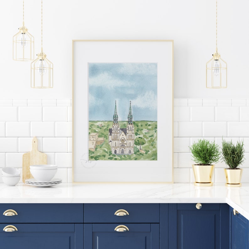 May include: A watercolor painting of a church with two tall spires. The church is painted in shades of white and gray, with green accents. The church is surrounded by green trees and bushes. The painting is framed in a light wood frame and is hanging on a white tiled wall. There are two gold pendant lights hanging above the painting.