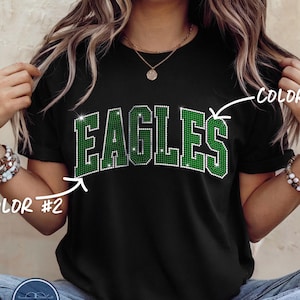May include: A black short-sleeved t-shirt featuring the word EAGLES in large, block-style letters. The letters are filled with green rhinestones and outlined in clear rhinestones. The text COLOR #1 and COLOR #2 with arrows point to the design.