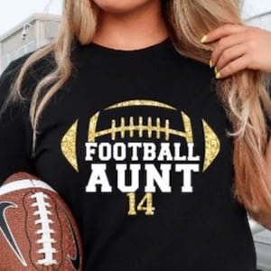 May include: Black t-shirt with gold glitter football design and the text "Football Aunt 14". A brown football is held in the left hand of the person wearing the shirt. A gold glitter jersey patch with the text "Williams 22" is in the lower right corner of the image.