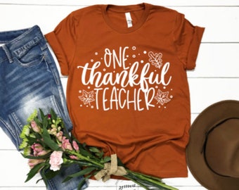 May include: A rust-colored t-shirt with the text "One Thankful Teacher" in white lettering. The shirt has a fall theme with white leaf designs.