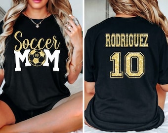 Peut inclure: T-shirt noir avec un texte en paillettes dorées qui dit "Soccer Mom" et un ballon de football avec le numéro 10 dessus. Le dos du t-shirt a un texte en paillettes dorées qui dit "Rodriguez" et le numéro 10.