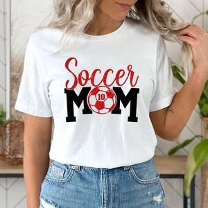 Op de afbeelding: Wit T-shirt met rode glittertekst "Soccer Mom" en een rode en witte voetbal met nummer 10.