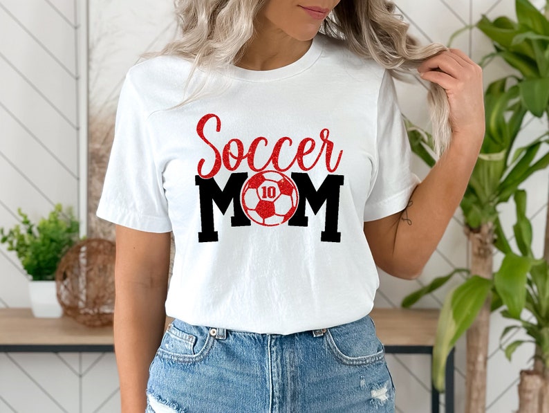 Op de afbeelding: Wit T-shirt met rode glittertekst "Soccer Mom" en een rode en witte voetbal met nummer 10.