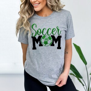 Op de afbeelding: Grijs T-shirt met de tekst "Soccer Mom" in zwart en groene glitter. Een groene voetbal met een witte "10" bevindt zich in het midden van de tekst.