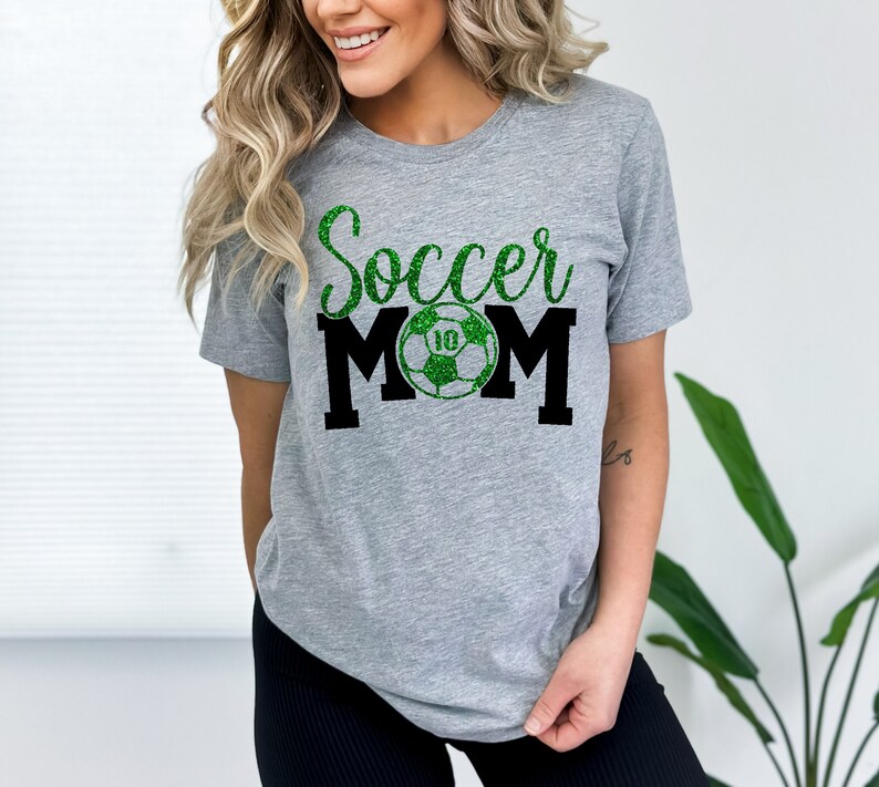 Op de afbeelding: Grijs T-shirt met de tekst "Soccer Mom" in zwart en groene glitter. Een groene voetbal met een witte "10" bevindt zich in het midden van de tekst.