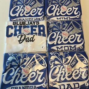 May include: Six blue and white t-shirts with the words "Cheer" and "Her Whole Heart" on them. The shirts are for different family members: Grandma, Mimi, Dad, Mom, Grandpa, and Dad. The shirts are packaged individually.