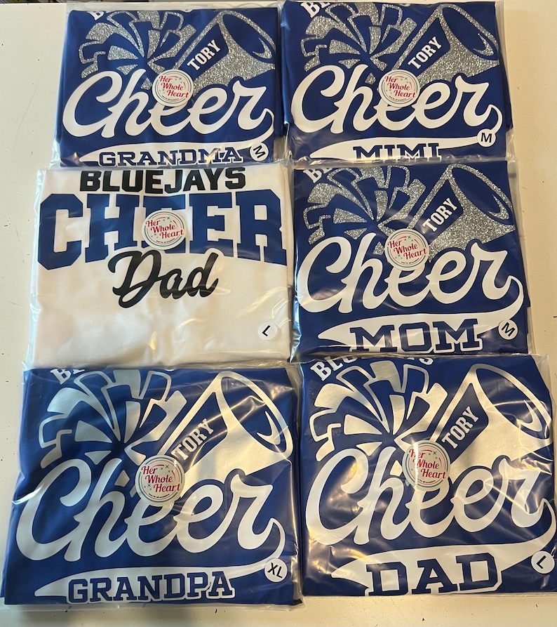 May include: Six blue and white t-shirts with the words "Cheer" and "Her Whole Heart" on them. The shirts are for different family members: Grandma, Mimi, Dad, Mom, Grandpa, and Dad. The shirts are packaged individually.