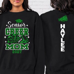 May include: A black long-sleeve sweatshirt featuring a green glitter graphic on the front that reads Senior Cheer Mom 2027 with a megaphone and pom-pom. The back of the sweatshirt displays a green glitter megaphone and the name HAYLEE in white text.