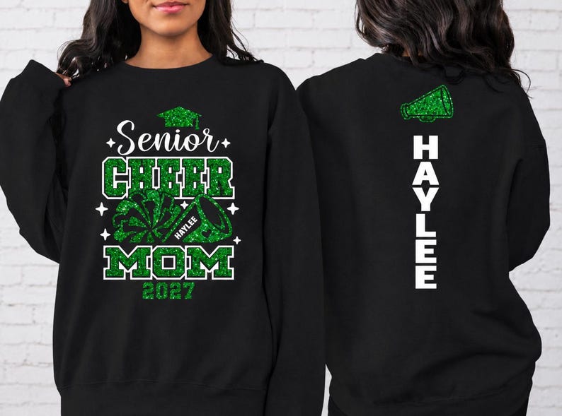 May include: A black long-sleeve sweatshirt featuring a green glitter graphic on the front that reads Senior Cheer Mom 2027 with a megaphone and pom-pom. The back of the sweatshirt displays a green glitter megaphone and the name HAYLEE in white text.