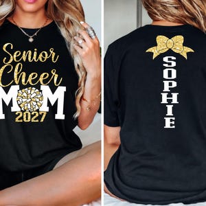 May include: Black t-shirt with gold glitter text reading "Senior Cheer Mom 2027" on the front. The back of the shirt has a gold glitter bow and the name "Sophie" in white. A cheerleading-themed apparel item.