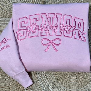 May include: A light pink sweatshirt featuring dark pink embroidery. The front displays the text SENIOR class of 2027 above a bow graphic. The sleeve is embroidered with a bow and the name Lanie in a cursive script font.