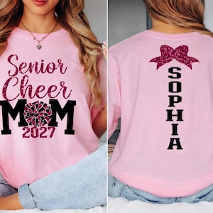 May include: Pink t-shirt with "Senior Cheer Mom 2027" text and a cheerleading graphic on the front. The back of the shirt features a bow graphic and the name "SOPHIA" in black. A comfortable, casual top.