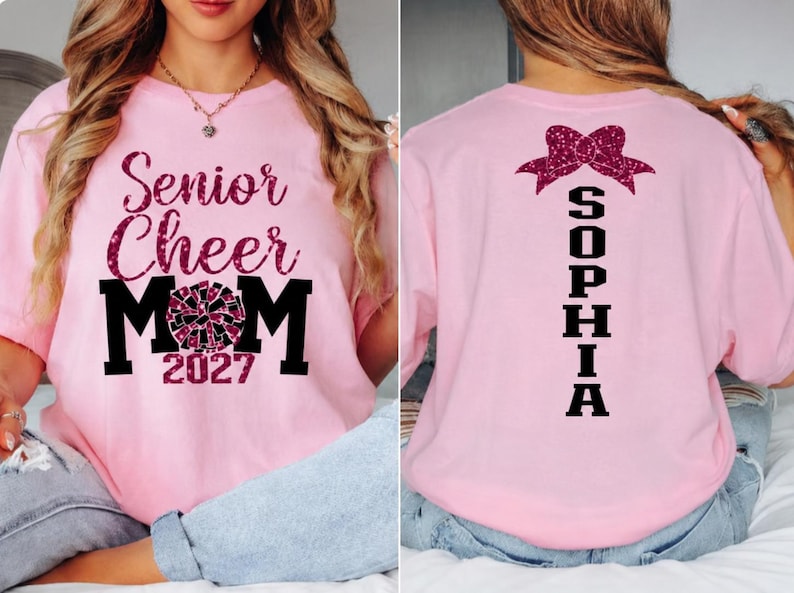 May include: Pink t-shirt with "Senior Cheer Mom 2027" text and a cheerleading graphic on the front. The back of the shirt features a bow graphic and the name "SOPHIA" in black. A comfortable, casual top.