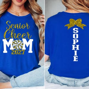 May include: Royal blue t-shirt with gold glitter text reading "Senior Cheer Mom 2027" and a cheerleading pom-pom graphic. The back of the shirt features a gold bow and the name "Sophie" in white.