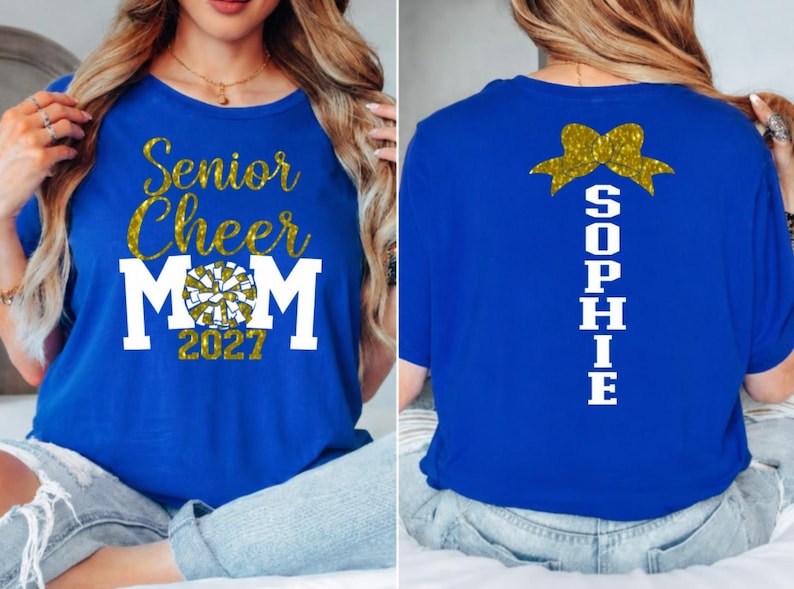 May include: Royal blue t-shirt with gold glitter text reading "Senior Cheer Mom 2027" and a cheerleading pom-pom graphic. The back of the shirt features a gold bow and the name "Sophie" in white.