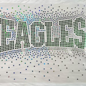 May include: A heat transfer decal featuring the word EAGLES spelled out in dark green rhinestones. The letters are outlined with iridescent, light-reflecting crystals that scatter into a gradient pattern of smaller dots across the white background.