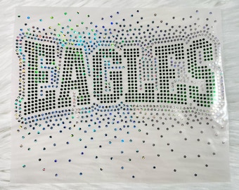 May include: A heat transfer decal featuring the word EAGLES spelled out in dark green rhinestones. The letters are outlined with iridescent, light-reflecting crystals that scatter into a gradient pattern of smaller dots across the white background.