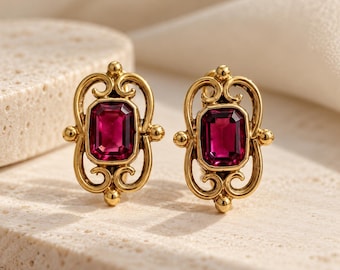 May include: A pair of gold-toned earrings featuring a central rectangular deep pink gemstone. The metal setting is crafted with intricate swirling scrollwork patterns surrounding the stone, with small spherical accents at the top and bottom.