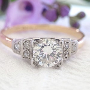 May include: A vintage engagement ring with a large, round, brilliant-cut diamond set in a platinum band. The band is accented with smaller diamonds on either side of the center stone.